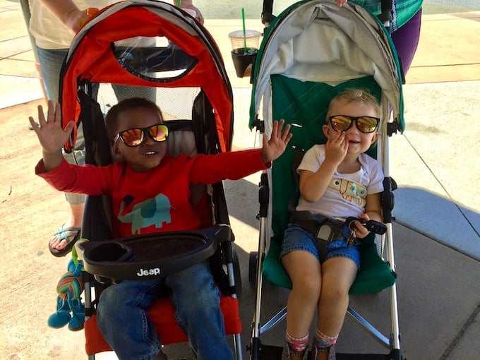 two young children sit in side-by-side strollers, wearing sunglasses and smiling