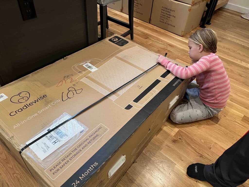 A large Cradlewise box rests on a hardwood floor, where an elementary-aged girl works to remove tape