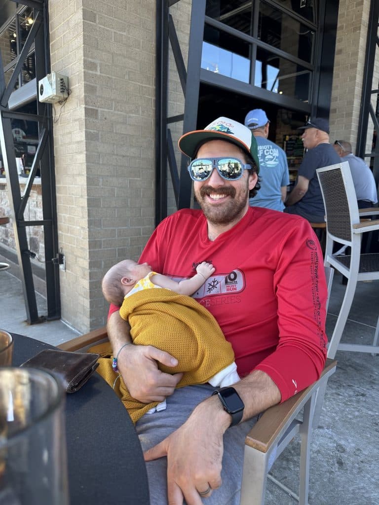 A dad sits at a restaurant holding his sleeping newborn. He's smiling at the camera.