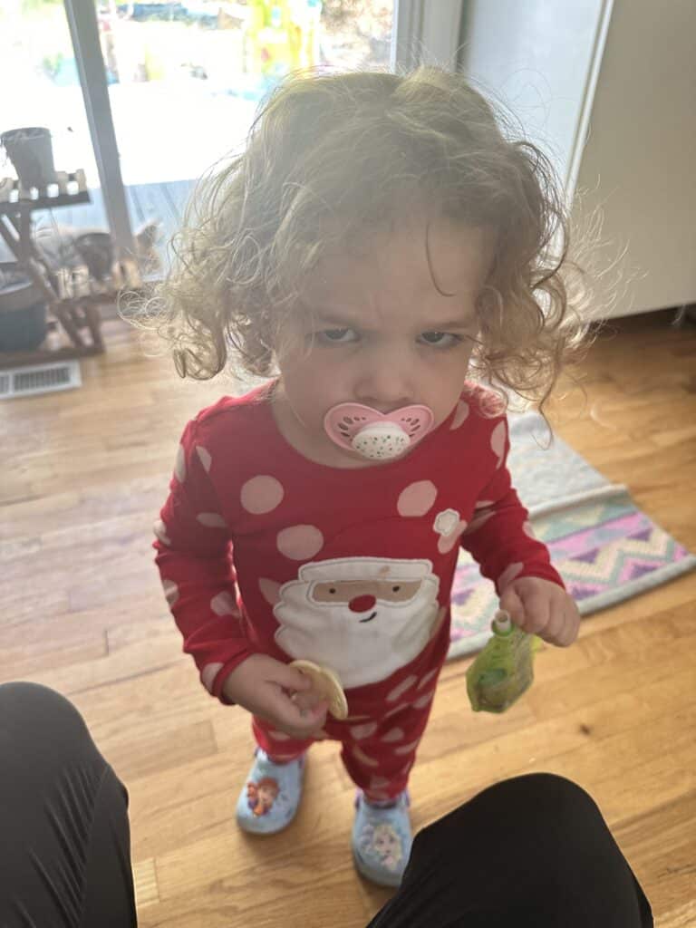 A toddler sucks on a pacifier and holds a back-up pacifier in her hand. She appears grumpy.