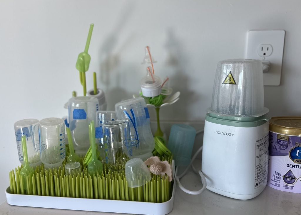 The Boon grass drying rack sits on a countertop, with six bottles and all of the bottle parts drying on top of it. The Momcozy bottle warmer and a canister of formula are also in the shot. 