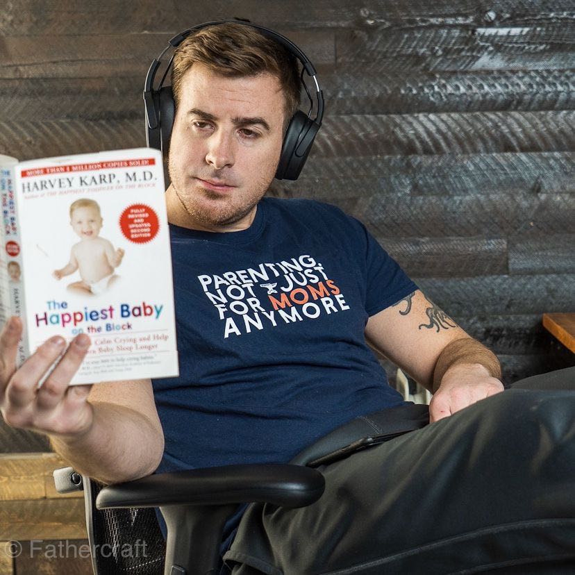 A guy reading a book with headphones wearing a parenting, not just for moms anymore t-shirt