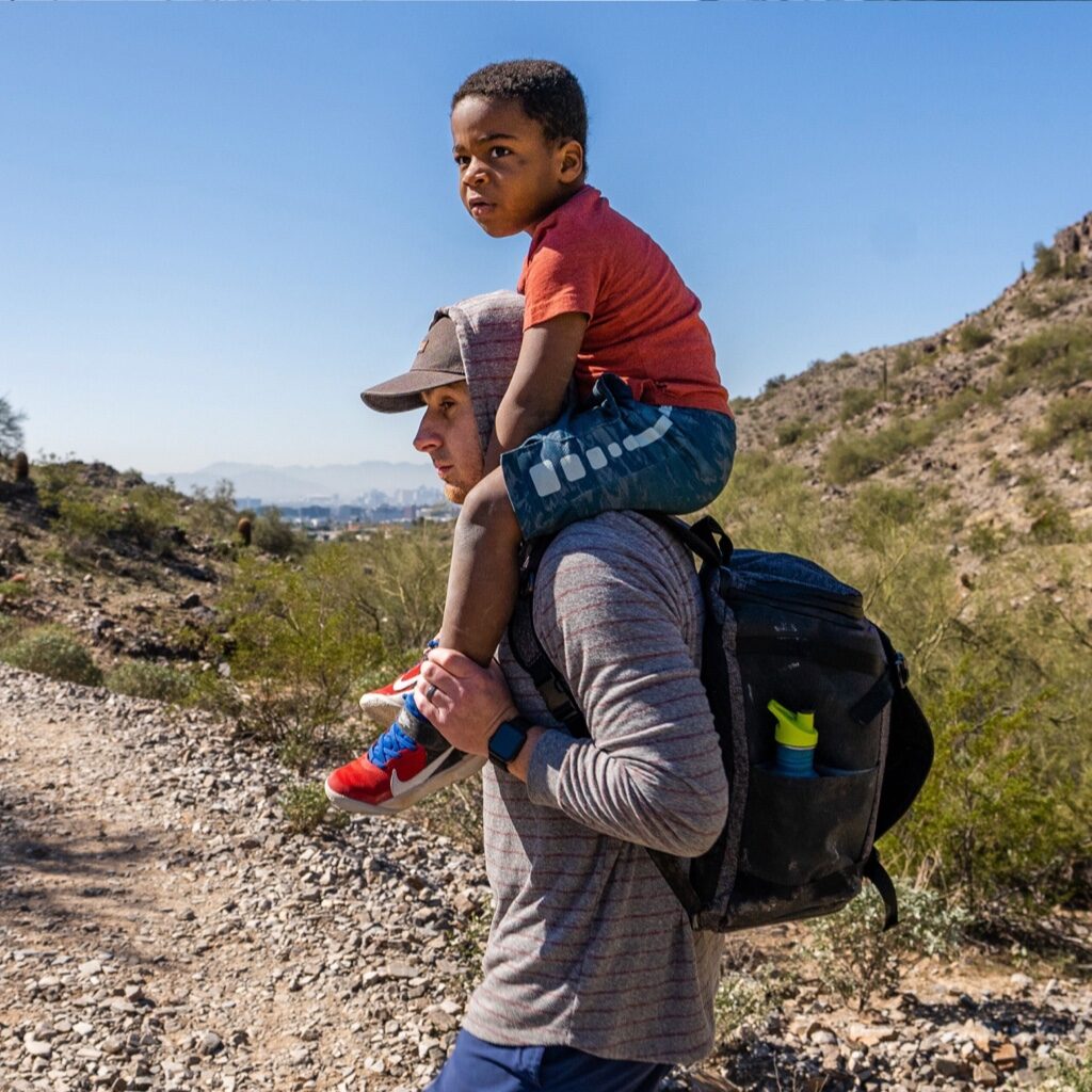 A dad with his son on his shoulders on a hike, carrying the Fathercraft backpack