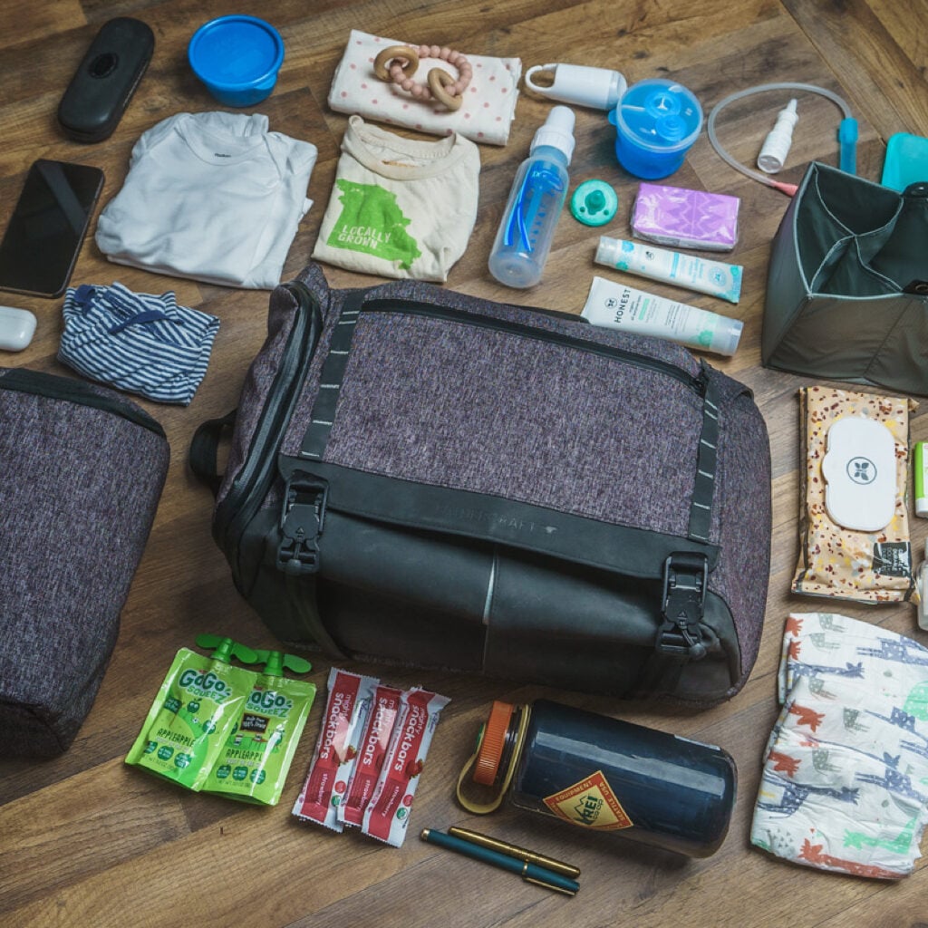 The Fathercraft diaper bag surrounded by all the items someone fits inside it, including snacks, diapers, a full-sized water bottle, a change of clothes, diaper cream, toys, Nose Frida, phone, pens, wipes, and bottles