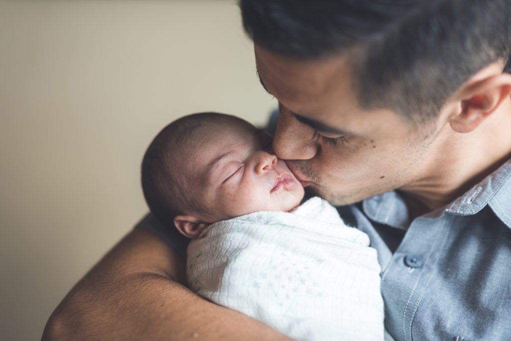 A dad gently kisses his newborn daughter on the cheek. He is holding her cradled in his arms as she is swaddled up comfortably for her nap.