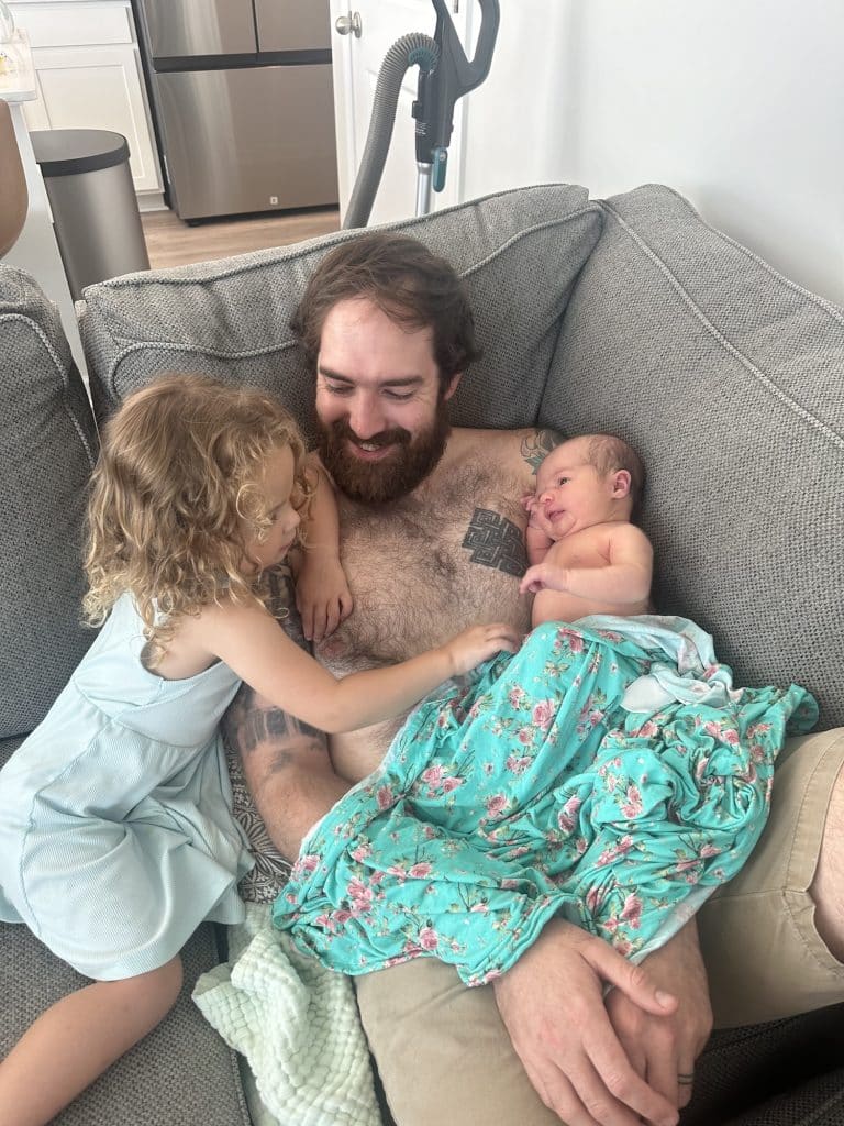 A dad does skin-to-skin contact with a newborn while his toddler plays with the baby
