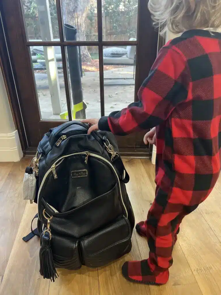 Black Itzy Ritzy backpack next to a toddler in red pajamas