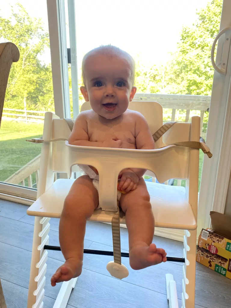 An infant sits in a white Stokke Tripp Trapp, smiling at the camera. In the image, there is no high chair tray, and the footplate has been removed. 